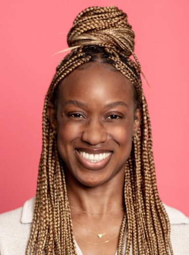 A woman with braided hair styled in a top bun smiles in front of a pink background. She is wearing a light-colored shirt and a small necklace.